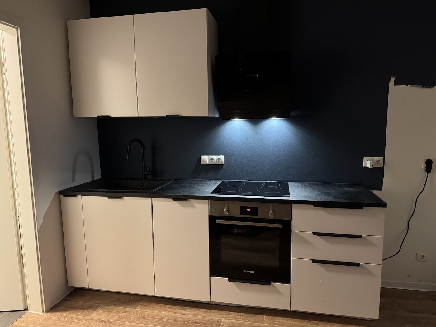 Fertige Küche mit weißen Fronten und Unterbauleuchten vor der dunklen Wand